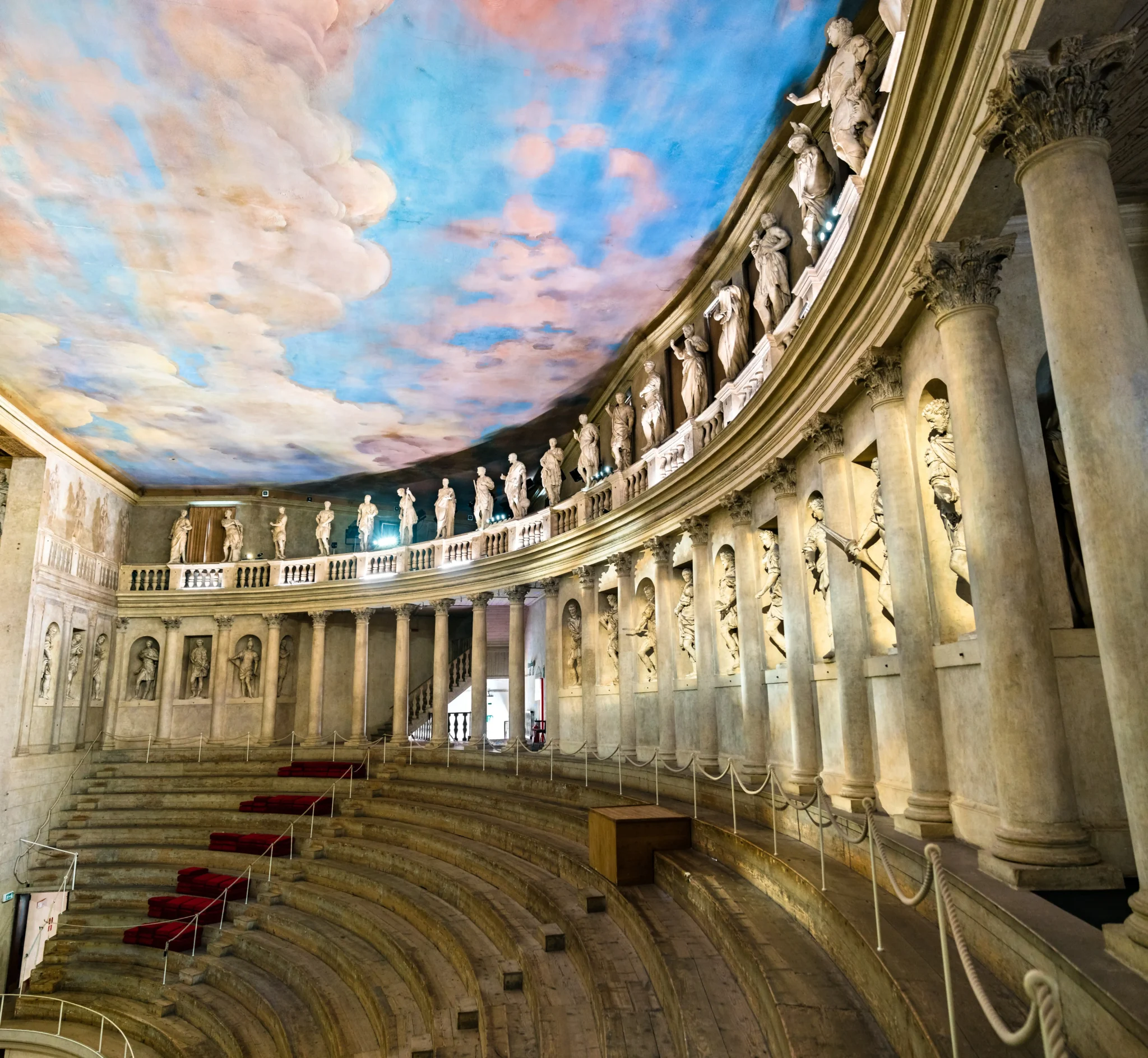 Teatro antico con gradinate in marmo e soffitto affrescato con cielo nuvoloso.