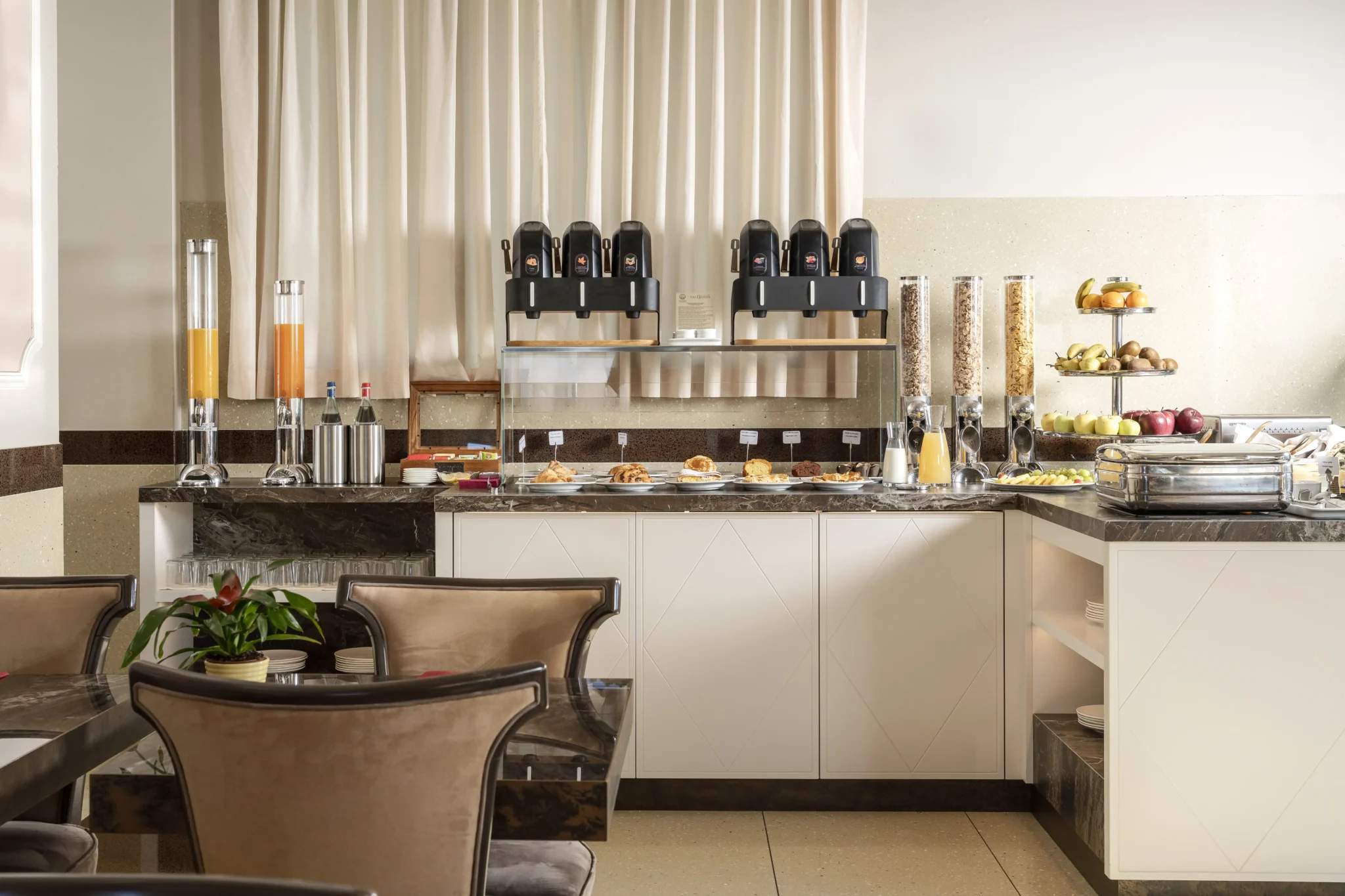 Area colazione di un hotel con buffet assortito di bevande, frutta e dolci.