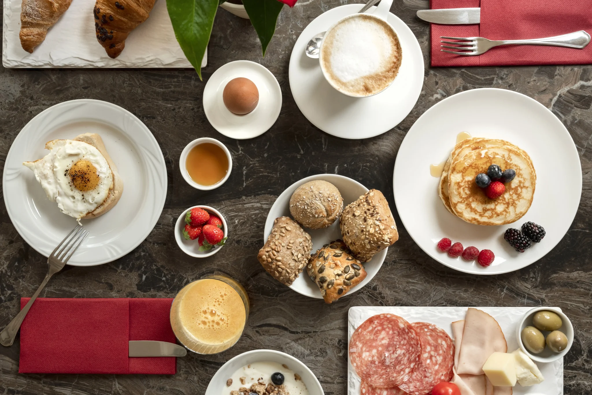 Colazione variegata con caffè, pancake, uova, pane, salumi e frutta.