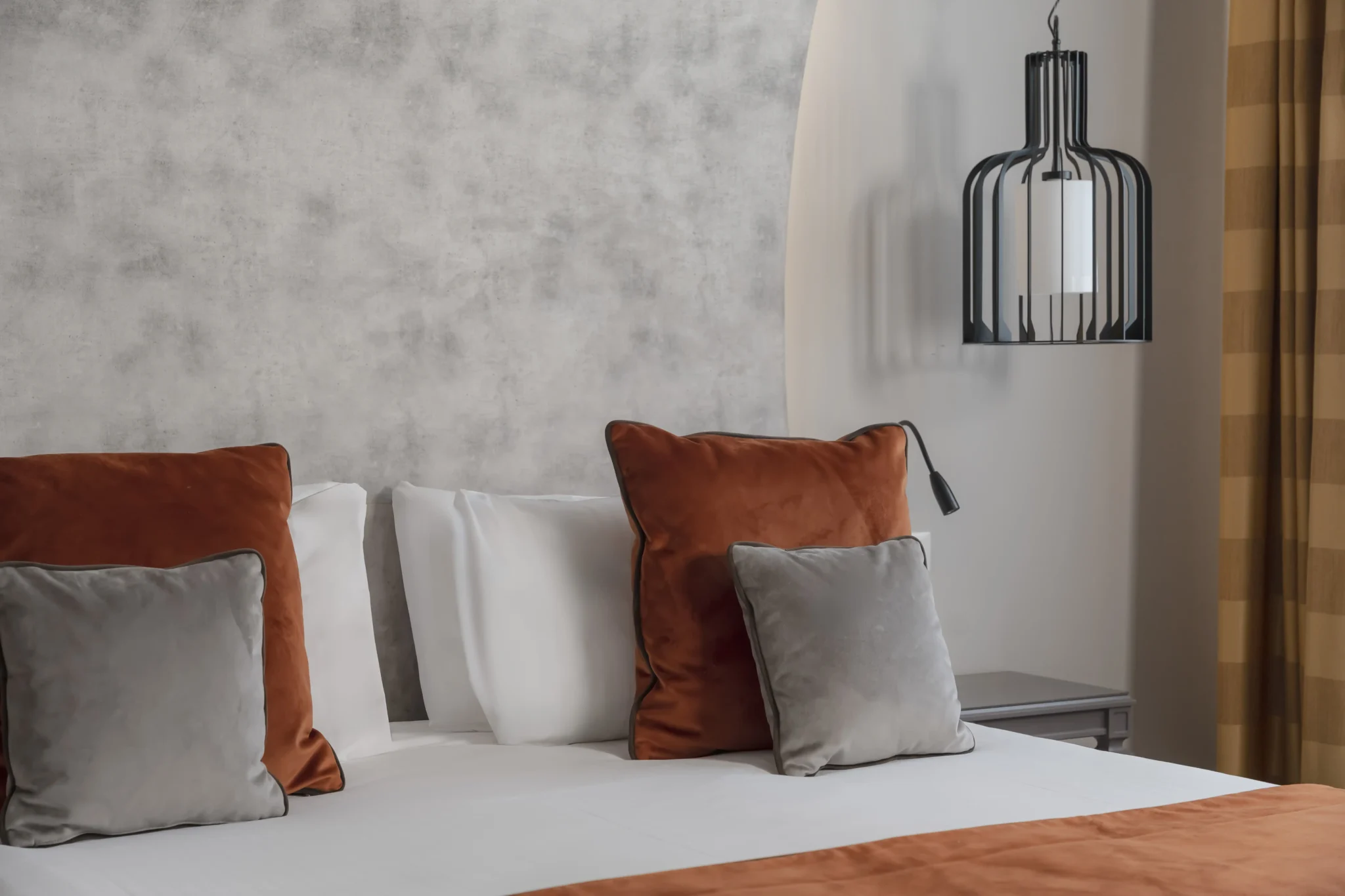 Letto moderno con cuscini arancioni e grigi, affiancato da una lampada sospesa.