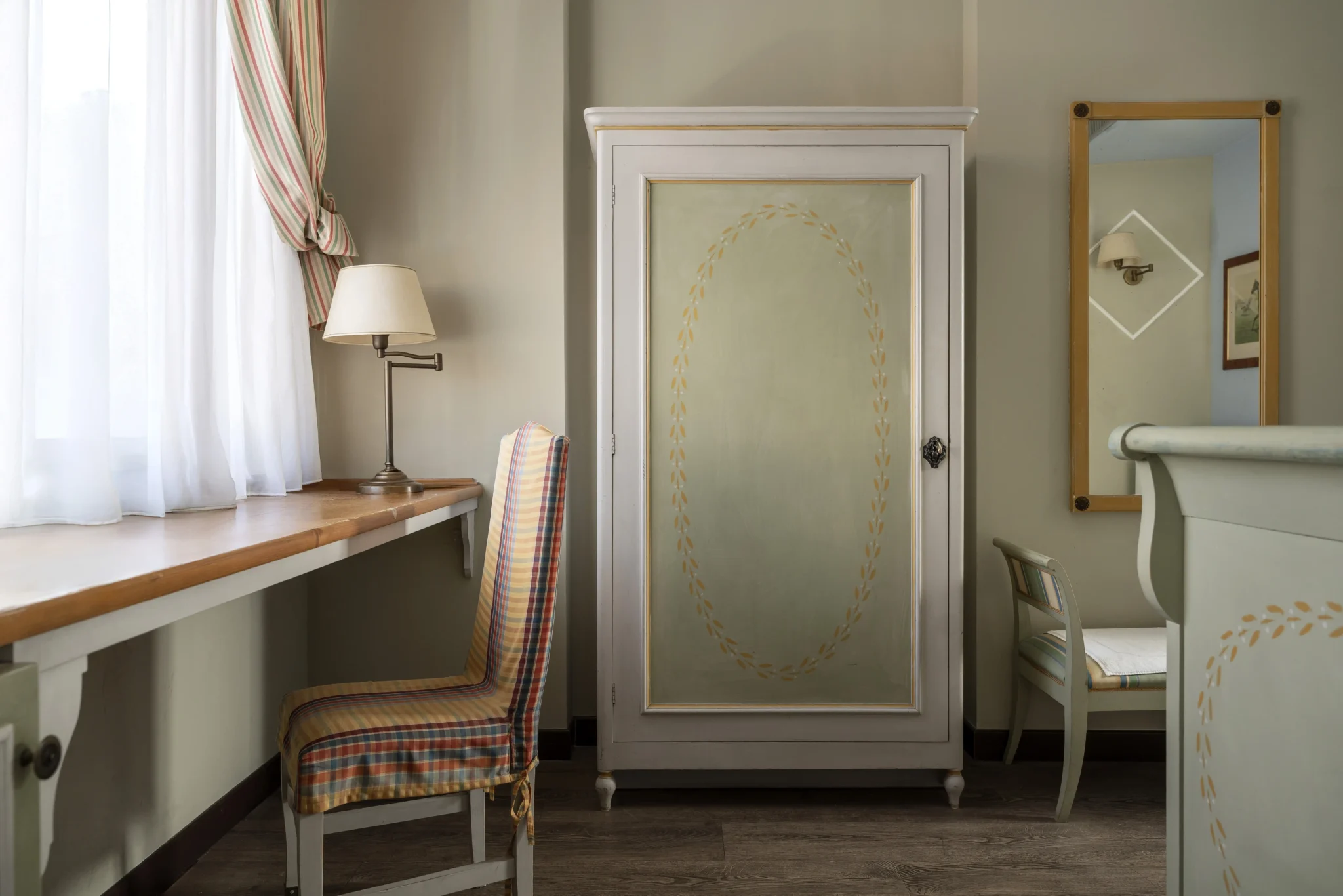 Interno di una stanza con arredo classico, include una scrivania, una sedia con copertura a quadri, un armadio dipinto e uno specchio.
