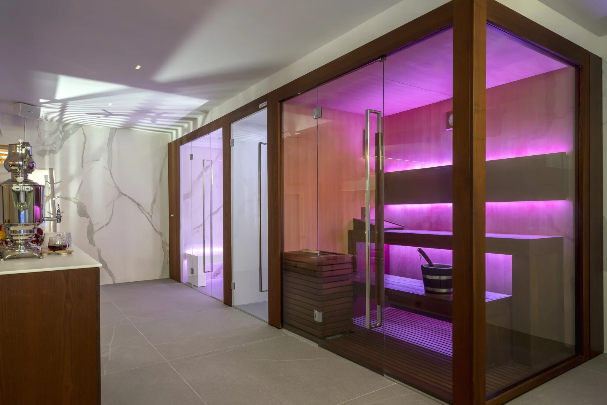 Vista di una moderna sauna illuminata con luce rosa accanto a una sala relax con macchina per bevande calde.