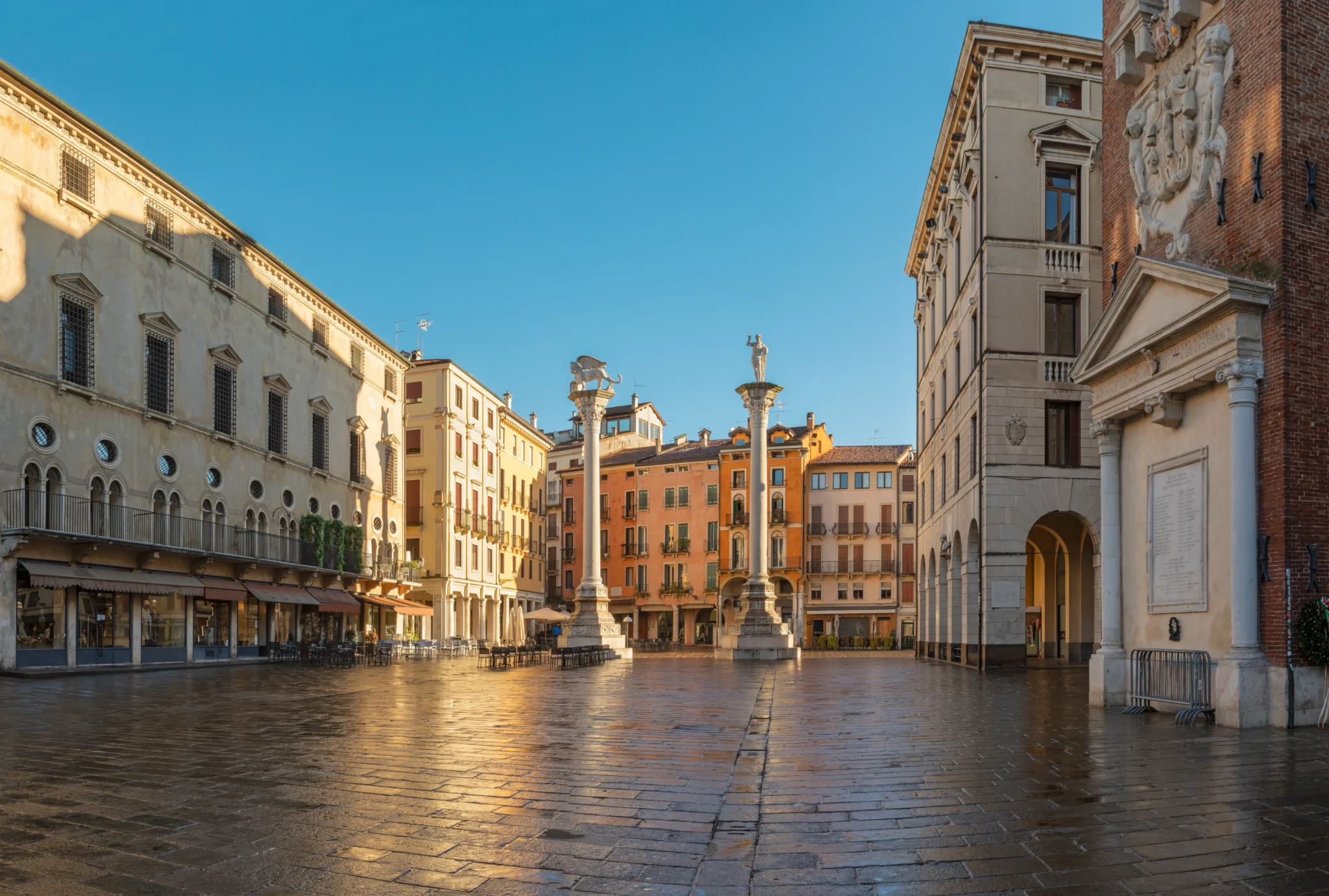 Vista di una piazza storica con colonne e edifici colorati illuminati dal sole.